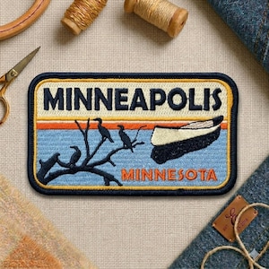May include: Embroidered patch featuring the city name "MINNEAPOLIS" above a scenic design with a boat, birds, and the state name "MINNESOTA". The patch has a navy blue border, with orange and light blue accents.