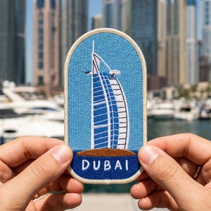 May include: An oval-shaped embroidered patch featuring a light blue background with a white and blue depiction of the Burj Al Arab building. Below the building is a brown island shape and the white text DUBAI, all bordered by a cream-colored edge.