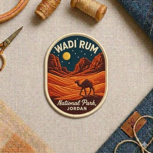 May include: An oval embroidered patch featuring a desert landscape with orange sand dunes, rocky mountains, a dark blue night sky with a yellow moon and stars, and a camel. The text reads WADI RUM NATIONAL PARK, JORDAN in white lettering.
