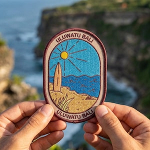 May include: Embroidered patch with a beach scene featuring a surfboard, sun, and ocean. The patch has a blue and yellow color scheme with the text "ULUATU BALI" in a circular design. The patch is held by hands.