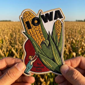 May include: An embroidered patch featuring two yellow corn cobs with green husks against a white background. The word IOWA is stitched in black letters at the top. A small figure in a hat and blue overalls stands on a red base with the text The Hawkeye State.