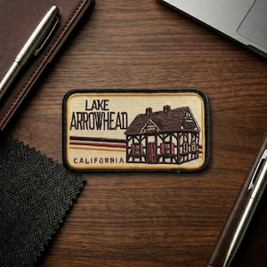 May include: Embroidered patch featuring a detailed illustration of a cabin with the text "Lake Arrowhead California". The patch has a tan background with brown, red, and black accents. The cabin has a brown roof and red doors.