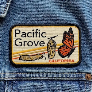 May include: A rectangular patch with the words "Pacific Grove CALIFORNIA" and illustrations of a caterpillar, chrysalis, and monarch butterfly. The patch has a cream background with orange and yellow stripes and is set against a blue denim jacket.