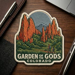 May include: Embroidered patch featuring a scenic view of the Garden of the Gods in Colorado. The patch depicts red rock formations, green foliage, and two hikers. The text "GARDEN OF THE GODS COLORADO" is displayed in a green border.
