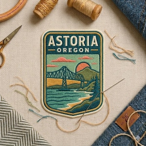 May include: A rectangular embroidered patch featuring the text ASTORIA OREGON. The design depicts a bridge spanning a body of water with a sunset, mountains, and trees in the background. The patch has a cream border and is set against a light fabric.