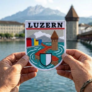 May include: A rectangular embroidered patch featuring the text LUZERN in black letters. The design depicts a brown tower, a red bridge, colorful buildings, and a white and green shield set against a backdrop of grey mountains and blue water.