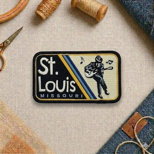 May include: Embroidered patch featuring "St. Louis Missouri" in white text on a black background, with a musician playing a guitar. The patch has a yellow and blue stripe design and a black border. Musical notes are also included.