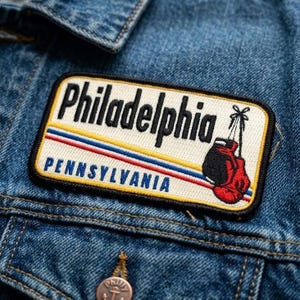 May include: A rectangular patch with the word "Philadelphia" in black letters, above "Pennsylvania" in blue. The patch features a red and black boxing glove design. The patch is sewn onto a blue denim jacket.