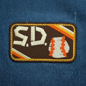 May include: Embroidered patch featuring the letters "S.D." in white, with a baseball design. The patch has a brown background with orange and yellow stripes, and a white baseball with orange stitching. The patch is rectangular with a yellow border.