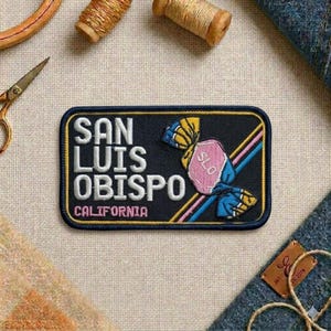 May include: Embroidered patch featuring the text "SAN LUIS OBISPO CALIFORNIA" in white lettering on a navy blue background. A candy graphic with pink and blue accents is on the right side. The patch is rectangular with a black border.