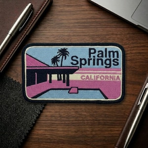 May include: A rectangular embroidered patch featuring a retro design with the words "Palm Springs CALIFORNIA". The patch has a blue background with a pink and black building and palm trees. The patch is surrounded by a black border.