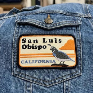 May include: A denim jacket with a patch featuring the text "San Luis Obispo CALIFORNIA". The patch has an illustration of a bird and an orange cloud design. The jacket is blue.
