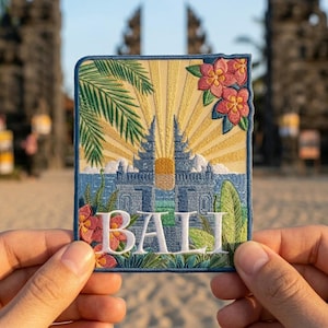 May include: A rectangular embroidered patch featuring a blue Balinese temple gate, a yellow sunburst, green palm fronds, and pink tropical flowers. The word BALI is stitched in white capital letters across the bottom center of the design.