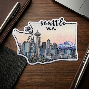 May include: Embroidered patch featuring a colorful depiction of the Seattle skyline, including the Space Needle and a mountain range. The patch is in the shape of Washington state with the text "Seattle W.A."