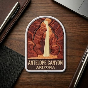 May include: Embroidered patch featuring a depiction of Antelope Canyon in Arizona. The patch has a brown border and a white arched top. The image shows the canyon walls in shades of brown and orange, with a light-colored waterfall. The text "ANTELOPE CANYON ARIZONA" is at the bottom.