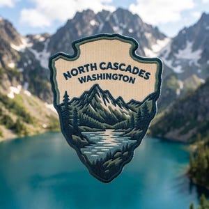 May include: A shield-shaped embroidered patch featuring a cream-colored background with the text NORTH CASCADES WASHINGTON in dark blue. The lower portion depicts a detailed landscape of dark blue and green mountains, pine trees, and a lake.