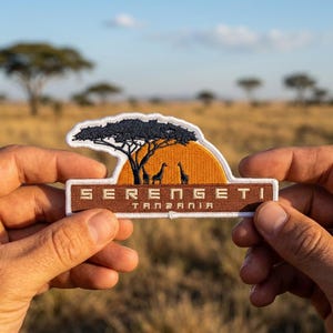 May include: An embroidered patch featuring a dark blue acacia tree silhouette against a bright orange sun, with two giraffes standing below. The brown rectangular base displays the text SERENGETI TANZANIA in white capital letters.