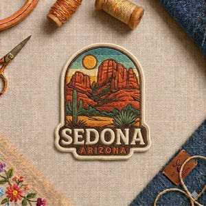 May include: An embroidered patch featuring a desert landscape with red rock formations, a yellow sun, and green cacti. The text SEDONA ARIZONA is stitched in cream-colored thread at the bottom of the arched design, which has a cream-colored border.