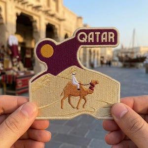 May include: An embroidered patch featuring a desert scene with a camel and rider in tan and brown threads. Above the dunes, a yellow sun sits against a deep purple sky. The word QATAR is stitched in white letters on a purple banner at the top.
