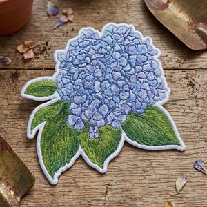 May include: An embroidered patch featuring a cluster of light blue and periwinkle hydrangea flowers with detailed green leaves. The design has a white border and is crafted with dense thread work to create a textured, realistic floral appearance.