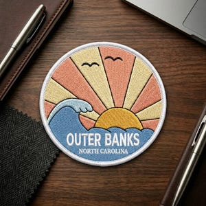 May include: A circular embroidered patch featuring a stylized sun with alternating yellow and peach rays, a blue ocean wave, and two black bird silhouettes. The text reads OUTER BANKS NORTH CAROLINA in white capital letters at the bottom.