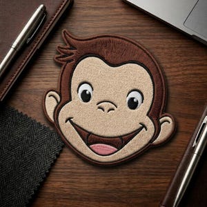 May include: Embroidered patch featuring the smiling face of a cartoon monkey with brown fur and a tan face. The monkey has large, expressive eyes and a wide, open mouth. The patch is likely for clothing or accessories.