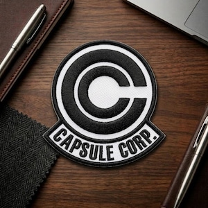 May include: Embroidered patch featuring the Capsule Corp logo from Dragon Ball. The patch is circular with a white center and a black outline. The text "CAPSULE CORP." is displayed in white on a black banner at the bottom.