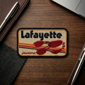 May include: Embroidered patch featuring the word "Lafayette" in black, with a red crawfish design and the word "Louisiana" below. The patch has a tan background with orange and brown stripes.