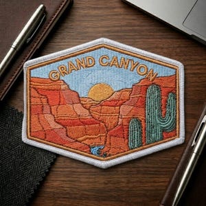 May include: Embroidered patch featuring a Grand Canyon landscape with a blue sky, orange canyon walls, a setting sun, and green cacti. The patch has a white border and the words "GRAND CANYON" in a gold color.