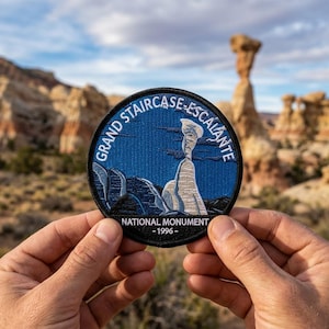 May include: A round patch featuring a landscape scene with the text "GRAND STAIRCASE-ESCALANTE NATIONAL MONUMENT - 1996." The patch depicts a rock formation against a blue sky. The patch is held in front of a blurred background of similar rock formations.