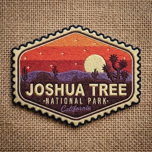 May include: An embroidered patch shaped like a postage stamp featuring a desert landscape at sunset with a yellow sun, purple mountains, and Joshua trees. The text reads JOSHUA TREE NATIONAL PARK California in cream-colored lettering.