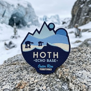 May include: A navy blue embroidered patch featuring a mountain landscape with two AT-AT walkers. The patch reads "HOTH - ECHO BASE - Outer Rim TERRITORIES" in white and light blue lettering. The patch is set against a snowy, mountainous backdrop.