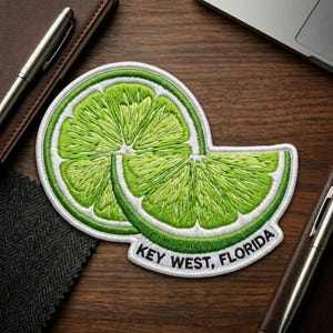 May include: A circular embroidered patch featuring a vibrant green lime slice design with a smaller lime wedge overlapping the front. The white border contains the black embroidered text KEY WEST, FLORIDA along the bottom edge of the patch.