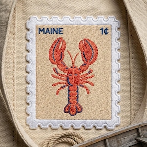 May include: A rectangular embroidered patch on a beige canvas fabric featuring a postage stamp design. The patch has a white scalloped border, the text MAINE in blue, the value 1 cent, and a central red lobster with blue detailing.