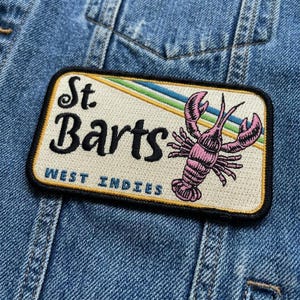 May include: Embroidered patch featuring the text "St. Barts WEST INDIES" with a pink lobster illustration. The patch has a cream background with a diagonal stripe of green, blue, and yellow. The patch is rectangular with a black border.