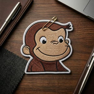 May include: Embroidered patch featuring a smiling monkey with brown fur and a white border. A gold safety pin is attached to the top of the patch. The patch is on a wooden surface, with pens and a notebook in the background.