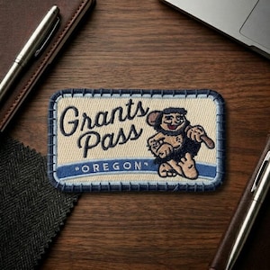 May include: Embroidered patch featuring the words "Grants Pass" and "OREGON" with a cartoon character holding a club. The patch has a cream-colored background with blue and black stitching.