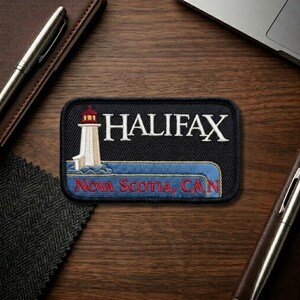 May include: A rectangular black patch featuring the word "HALIFAX" in white, above an illustration of a lighthouse. Below the lighthouse is the text "Nova Scotia, CAN" in red. The patch has a blue border.