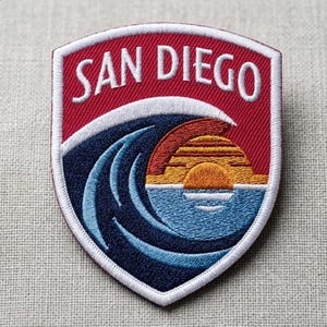 May include: A shield-shaped embroidered patch featuring the text SAN DIEGO in white letters on a red background. Below the text, a dark blue ocean wave curls over a stylized orange and yellow sunset reflecting on light blue water.