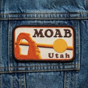 May include: Embroidered patch featuring the word "MOAB" above "Utah" with a desert landscape design. The patch includes an orange arch, a yellow sun, and brown and orange lines, all set against a cream background, sewn onto a denim jacket.
