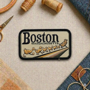 May include: A rectangular patch with the word "Boston" in dark letters, and "Massachusetts" below. The patch features a line of ducklings following a mother duck. The patch is bordered in black.