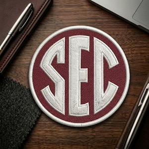May include: Embroidered patch featuring the letters "SEC" in white, set against a maroon background within a circular shape. The patch has a white border and is likely for apparel or accessories.