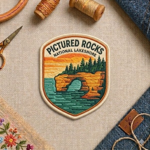 May include: A shield-shaped embroidered patch featuring a landscape of Pictured Rocks National Lakeshore. The design includes a yellow and orange sunset, a green forest, and a teal water archway. The text reads Pictured Rocks National Lakeshore.