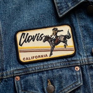 May include: A rectangular patch featuring a cowboy riding a bucking bronco, with the word "Clovis" in cursive and "CALIFORNIA" below. The patch has a cream background, black border, and is sewn onto a blue denim jacket.