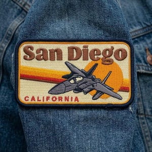 May include: A denim jacket with a rectangular patch featuring the words "San Diego" in brown lettering. The patch includes a gray jet plane, a yellow and orange sunset design, and the word "CALIFORNIA" in red.