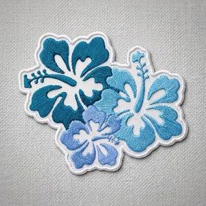 May include: An embroidered patch featuring three hibiscus flowers in varying shades of blue, ranging from dark teal to light sky blue. The design has a white border and is stitched with detailed thread work on a textured white fabric background.