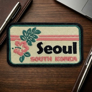 May include: Embroidered patch with the word "Seoul" in black, and "SOUTH KOREA" in pink. The patch features a floral design with pink and green accents, and a black border. The background is beige.