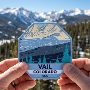 May include: A blue and white embroidered patch featuring a mountain landscape with the text "VAIL COLORADO USA". The patch has a geometric shape and is held up against a backdrop of a snowy mountain range.