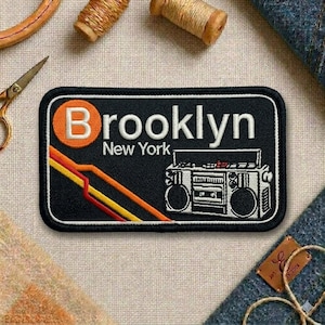 May include: A black embroidered patch featuring the word "Brooklyn" in white, with an orange "B" in a circle. Below, "New York" is written in white. The patch includes a retro boombox design and colorful stripes.