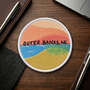 May include: A circular embroidered patch featuring a landscape design with layered peach and orange dunes, a blue water section, and yellow sand with green beach grass. The text OUTER BANKS, NC is stitched in black across the center of the patch.